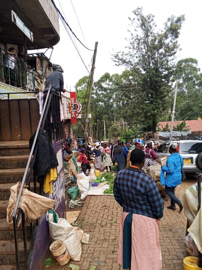 The image depicts an active outdoor market scene in Kisii Central ward, Kenya, viewed from a slightly elevated position. In the immediate foreground, the back of an individual wearing a blue and black plaid shirt and a multi-patterned skirt (red/white checkered and dark teal) is visible, facing away from the viewer. To the left, a set of brown concrete stairs with a black metal railing ascends towards a building. A black jacket and a yellow cloth hang over this railing. At the base of the stairs, several soiled burlap sacks and an empty white bucket with a red label are on the uneven, brown interlocking brick pavement, which shows scattered green debris.
The midground features a crowded market area. Numerous individuals, primarily adults, are visible, many engaged in commercial activity. Produce, including leafy green vegetables, is displayed on white tarps and directly on the ground. Various sacks and yellow plastic containers are interspersed among the market stalls. On the left, a multi-story building with a gray concrete overhang and exposed utilities is present. One person in a white shirt is visible on an upper-level balcony, while another, wearing a patterned top, leans over a lower railing. A prominent red sign with white text reading "SHIVLING" and an accompanying logo is affixed to this building. Overhead, multiple utility poles support a dense network of electrical and communication lines. Towards the right, a white SUV or similar vehicle is parked, displaying a visible Kenyan license plate, partially reading "KCQ 942C." A woman in a bright blue jacket, carrying a blue bag, walks rightwards past the vehicle.
The background consists of dense green foliage from numerous trees under an overcast sky. Distant structures with reddish-brown roofs are partially visible through the trees. The overall environment appears bustling and functional.