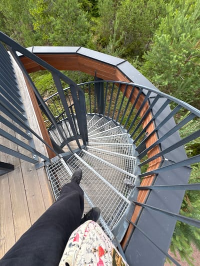 The image shows a view from the top of a spiral staircase, looking down. The staircase is made of metal mesh steps and has a dark grey metal railing. The staircase is surrounded by wooden decking and sections of wood paneling on the exterior. Below the staircase, there is dense green forest with pine trees visible. A person's legs, clad in black pants and dark sneakers, are visible at the bottom of the frame, suggesting they are standing on the wooden platform at the top of the stairs. A patterned bag rests on their lap. The overall scene depicts an outdoor observation tower or viewpoint within a forested area. The location is identified as Peski, Russia.