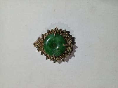 A single piece of jewelry, possibly a brooch or pendant, is centered on a plain, light grey background. The item consists of a prominent, polished, donut-shaped green stone, likely jade, featuring a central circular aperture. This green stone is held by four golden-toned prongs. Surrounding the central stone is an ornate, golden-toned metallic setting, from which approximately two dozen smaller, square-cut, clear or light-colored stones radiate outwards in a starburst or floral pattern. The object casts a distinct shadow to its lower left, indicating illumination from the upper right. No individuals, actions, or interactions are depicted. The image contains no visual information to determine its specific location within the city of Phakeo, Laos.