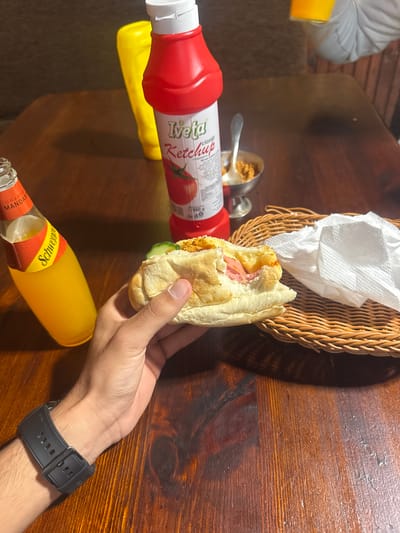 The image captures a casual meal setting on a wooden table, likely indoors in Konyavo, Bulgaria. The central focus is a person's right hand holding a half-eaten sandwich or hot dog. The sandwich has a light-colored bun and appears to contain a pink meat filling, possibly some melted cheese, and a slice of green vegetable like cucumber. A black smartwatch or fitness tracker, with "HUAWEI WATCH" faintly discernible on its strap, is worn on the person's wrist.
The table, made of dark, reddish-brown wood with visible grain, holds several items. A red squeeze bottle of "Ivela Ketchup Classic" is prominently displayed, lying horizontally. Behind it is a yellow squeeze bottle, likely containing mustard. To the upper right, a woven wicker basket holds several white, crumpled paper napkins. A small, metallic bowl containing a brownish, textured substance (possibly spices or crumbs) and a small spoon is visible near the ketchup bottle. In the lower part of the frame, a glass bottle of "Schweppes Mandarin" soda is lying down.
In the upper left corner, a portion of another person wearing a white garment can be seen, along with a glimpse of a yellow drink in a glass. The overall impression is a relaxed meal or snack.