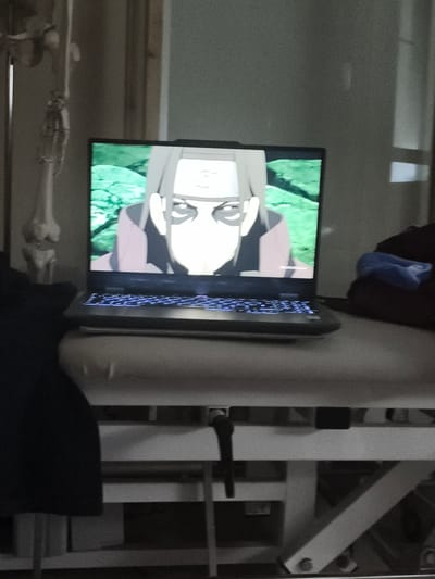 A laptop is open and displayed on a surface, showing a picture of an anime character with a headband and dark circles under its eyes. The character has light skin and is wearing a dark-colored garment with pink accents. The background of the image on the laptop is a blurred green landscape. The laptop's keyboard is illuminated with blue light. To the left of the laptop, a skeletal model of a human leg and foot is visible, hanging from a stand. The scene appears to be indoors, with a plain wall in the background. The laptop is placed on a light-colored, padded surface, which rests on a white metal frame structure that resembles medical equipment. A dark blue fabric is visible to the right of the laptop, partly covering a darker object. The image was taken in the city of Eitorf, Germany.