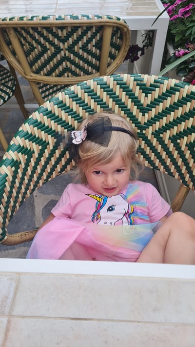 A young, fair-skinned girl with blonde hair is resting on a green and tan woven chair. She is wearing a light pink T-shirt with a unicorn graphic and a pink tulle skirt. A black headband with a black bow and a white butterfly adorns her hair. Other matching woven chairs are next to the chair the little girl is sitting on. There's a white table next to the chairs with purplish-pink flowers. The location appears to be outdoors, potentially in a courtyard, due to the light colored tile flooring. The location is in Kiryat Gat, Israel.