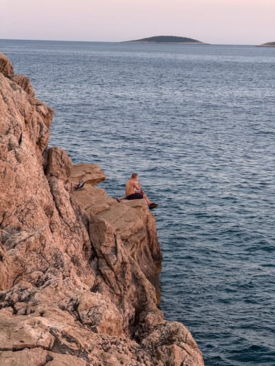 A young man, shirtless and wearing dark shorts, sits on the edge of a rocky cliff overlooking the Adriatic Sea in Primošten, Croatia. The cliff face, composed of rugged, reddish-brown rock, dominates the left side of the frame. The sea, a deep blue, stretches out to the horizon, where a few small, tree-covered islands are visible. The sky above is a pale, hazy pink, suggesting either dawn or dusk. The man appears to be holding a small object in his hands, and his gaze is directed out towards the water. His legs are casually crossed, with one foot resting on a lower ledge. The overall impression is one of peaceful solitude against a dramatic natural backdrop.