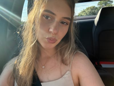 The media file shows a close-up, vertically oriented, selfie-style portrait of a young woman. She has long, blonde hair that is slightly windblown and catches the sunlight. She is wearing a thin-strapped top and a delicate necklace with a small heart-shaped pendant. Her face is turned towards the camera, with her eyes looking directly at the viewer. She has on subtle makeup, including eyeliner and lip gloss. The background is dark and appears to be the interior of a car, possibly a seat and a headrest. Sunlight is streaming in from the right side, creating lens flares and illuminating her hair. Outside the car window, through a slightly blurred view, a bright sky and green trees can be seen, suggesting it is daytime with fair weather. The overall mood is casual and relaxed, possibly taken while traveling. There is no visible text in the media file.