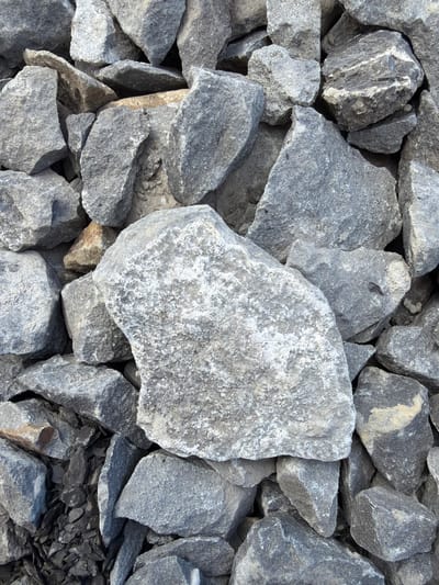 The image displays a close-up, overhead view of a pile of rough, gray stones. The stones vary in size and shape, with many having angular and irregular surfaces. Some stones exhibit lighter patches, suggesting wear or mineral deposits. The overall texture is coarse and natural. The background is composed of more of the same type of stones, creating a dense and textured scene. There is no indication of human presence or activity. The location is Jodłownik, Poland.