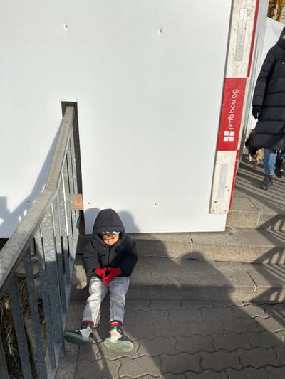A young child is seated on a concrete step in Neuhausen am Rheinfall, Switzerland. The child is wearing a black hooded puffer jacket, light grey trousers, red knitted gloves, and light-colored athletic shoes with translucent elements and green soles. A multi-colored striped headband is visible beneath the hood. Their hands are clasped together, and their head is bowed, looking downwards.
To the child's left, a metallic handrail runs diagonally upwards. The immediate ground consists of grey interlocking pavers. A tall, white corrugated construction barrier or temporary wall serves as the backdrop behind the child and the railing. On the right side of the image, a section of the white barrier features a vertical red support beam with "pmb bau ag" printed in white lettering, indicating a construction company.
Behind the barrier on the right, an adult figure, identifiable by a black puffer jacket and blue jeans, is partially visible, walking leftward. A leashed brown dog walks alongside the adult. Faintly in the upper right background, a yellow construction crane boom is visible above the barrier. The scene is brightly lit by direct sunlight, casting distinct, elongated shadows on the steps and paved ground.