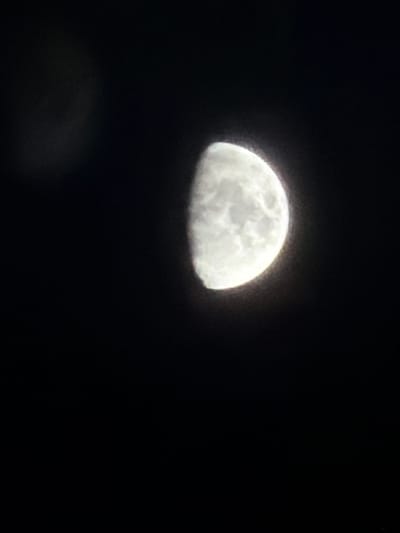 The media file is a photograph of a moon in a dark night sky. The moon is partially illuminated, showing surface details and craters. The overall impression is of a celestial event in a remote or rural setting, possibly a clear night due to the visibility of the moon. There are no people or any text visible in the image. The setting is outdoors, under the night sky.