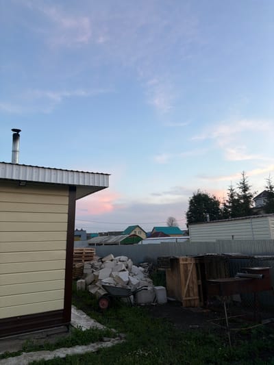 The image depicts a backyard scene in Nizhny Suyuk-Su, Russia, during what appears to be late afternoon or early evening, given the soft lighting and pastel hues in the sky. In the foreground to the left is the side of a light-colored, corrugated building with a dark brown trim along the base and corner. A metal chimney extends vertically from the roof of this building. Adjacent to this structure, a pile of light-colored rubble, possibly construction debris, is present, with a wheelbarrow to the left of the pile. Behind the rubble, a wooden shed with a slanted roof and a door made of vertical planks is visible. To the right of the shed, a rusty barbecue grill with two cylindrical pipes on top is set up on legs and enclosed by a chain-link fence. In the background, a series of houses with various colored roofs, including green and red, are scattered across the landscape. Pine trees are visible behind the houses. The sky is a soft blue with wispy, pink-tinged clouds. The ground is covered with grass and some concrete pathways or slabs. There are no people visible in the image.