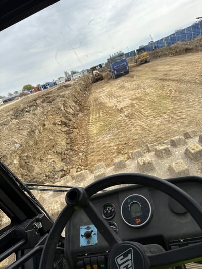 The image depicts the interior of a JCB vehicle, likely a backhoe loader, on a construction site near Hristiyanovo, Bulgaria. The steering wheel and dashboard are in the foreground, showing the odometer reading 249.9 hours. Through the front windshield, a barren, dirt-covered construction site is visible. Rows of blue shipping containers line the left side of the frame. Several construction vehicles, including trucks and possibly excavators, are parked along the containers. The sky is overcast.