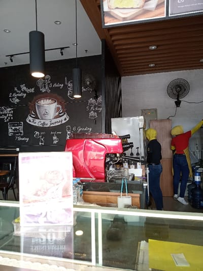 This image captures the interior of a coffee shop or cafe, located in Kecamatan Panji, Indonesia. The scene is dominated by a service counter featuring a prominent red espresso machine. Behind this counter, two individuals, likely staff members, are visible; one is standing facing away from the camera, wearing dark attire and a yellow head covering, while the other is reaching upwards, dressed in a red shirt, blue jeans, and a yellow head covering.
The left wall serves as a large chalkboard-style mural, depicting "The Coffee Journey" with illustrations and text outlining stages like "2. Harvesting," "3. Huling," "Roasting," "5. Packag," "6. Grinding," and "7. Brewing." A large coffee cup with latte art is central to the mural. Two dark cylindrical pendant lights hang from the ceiling, illuminating the area. A black wall-mounted fan is also visible on this wall. The right wall is light-colored, possibly brick, and features another black wall-mounted fan and a silver refrigerator.
Above the counter on the right, framed items are displayed, one showing toast with butter and another with text, including "Hurga par sotien Ribuc." The ceiling has a decorative wooden slatted design with recessed lighting. In the foreground, a glass display counter reflects the interior. A white stand with a purple-toned image and some indistinct text is visible on or within this display. Bottles of water, various syrup dispensers, and a large blue water jug on a dispenser are also present behind the main counter. The activity suggests a busy cafe setting, with staff attending to the service area. The overall impression is a modern, functional coffee preparation space.