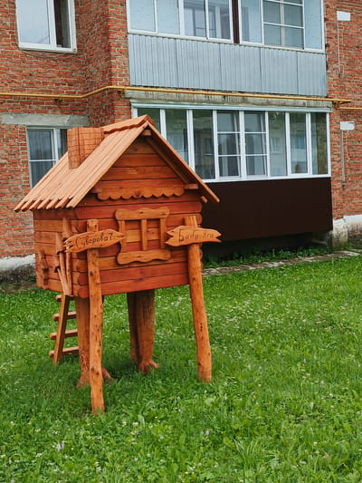 A quaint, rustic wooden structure resembling a small house sits on a grassy lawn. The house is elevated on four log-like supports, and a small ladder leans against its side. It features a charming, shingled roof and what appears to be a chimney made of layered wooden bricks. Two carved signs are attached to a central wooden post in front of the house. The sign on the left, pointing left, has Cyrillic text that reads "ул. Суворова" (Suvorova St.). The sign on the right, pointing right, has Cyrillic text that reads "Баба Яга" (Baba Yaga).

Behind the wooden house, a multi-story brick building stands. It has several windows, some with white frames and some with silver paneling, suggesting a balcony or enclosed porch area. A yellow pipe runs horizontally along the side of the building. The overall scene is set outdoors on a bright, perhaps sunny day, as indicated by the well-lit environment and vibrant green grass dotted with small white flowers. The presence of the Baba Yaga sign hints at a playful, perhaps fairytale-themed, decoration or attraction. The location is identified as Medvedyovka, Russia.