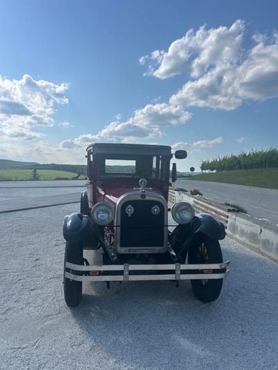An antique red car is parked on a gravel surface in Ulmu, Moldova. The car is facing forward and is the central subject of the image. The background features a paved pathway, a low stone wall with wooden accents, a vineyard on a hillside, and rolling green hills under a blue sky with scattered white clouds. The lighting suggests it is daytime, likely late afternoon or early evening. There are no people visible in the image.