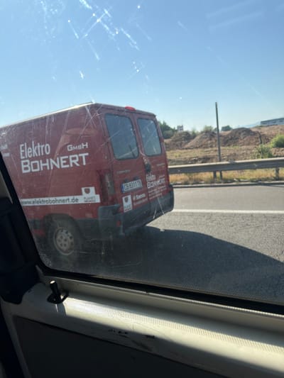 An exterior medium shot captures a red van with white lettering parked on the side of a road. The van is labeled "Elektro BOHNERT GMBH" and has a website address "www.elektrobohnert.de". A license plate reading "CB 8311 TB" is visible on the back of the van. The van appears to be a Citroen. The background shows a construction site with piles of dirt and a few trees. The sky is clear and blue, with some lens flare visible. The image is taken from inside another vehicle, with a window and a door handle in the foreground. The scene is set in Dolni Bogrov, Bulgaria.