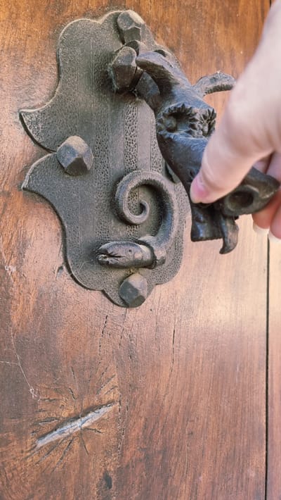A close-up, eye-level shot captures a hand reaching for an ornate, dark metal door knocker on a rustic wooden door in Rupit, Spain. The door knocker is fashioned in the shape of a dragon or mythical creature, with its body intricately coiled and forming a snake-like head with an open mouth. The hand, belonging to a person with fair skin and short fingernails, is positioned to grasp the head of the creature. The wooden door exhibits a weathered texture with visible grain and a distinct knot with radiating cracks, suggesting age and character. The lighting is natural and casts subtle shadows, highlighting the detailed craftsmanship of the knocker and the texture of the wood.
