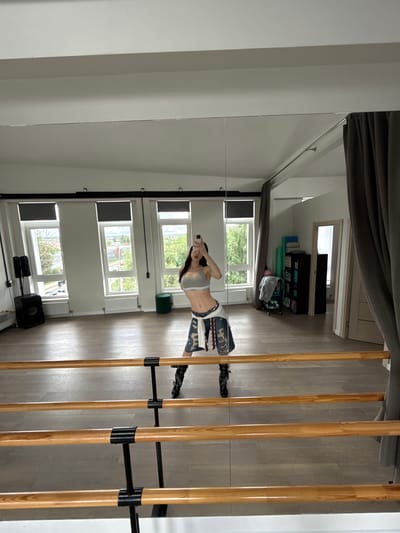 The image is a reflection in a large mirror, showing a young woman taking a selfie. She is wearing a grey crop top and denim shorts, with a white garment draped over her shoulder. She is standing in what appears to be a dance studio or gym, evidenced by the barre along the right side of the frame and yoga mats visible in the background.
The room has light-colored wooden floors and large windows that offer a view of green trees and buildings outside, suggesting it is daytime and likely spring or summer. The windows are covered with blinds, one of which is partially lowered. In the background, there is a black storage unit with various items on shelves, including what look like exercise balls and water bottles. A green yoga bolster is on the floor near the windows.
The woman's pose suggests she is actively taking a photograph of herself. Her expression is neutral, and the overall mood of the image is casual and everyday. The reflection distorts the perspective slightly, as is common with mirrored surfaces. The scene is well-lit, likely by natural light coming through the windows. The background elements, such as the wooden floor and the barre, are clear and in focus within the reflection. The image is oriented vertically, with the top of the scene appearing at the bottom due to the reflection. There is no visible text in the image.