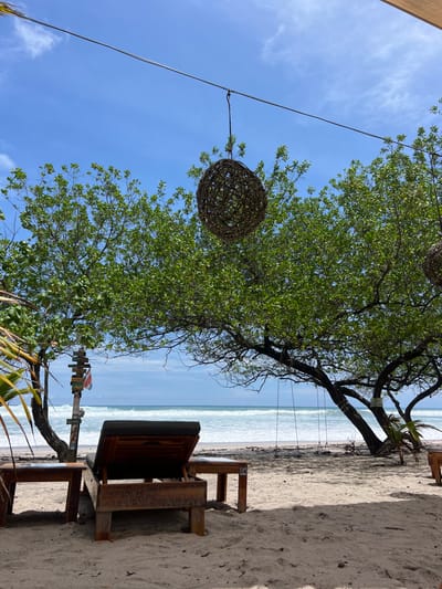 The image shows a beach scene in Santa Teresa, Costa Rica, with a large tree casting shade over a beachside lounge chair. The chair is made of wood and has a dark gray fabric cushion. In front of the chair are two small wooden tables. The beach is sandy and appears to be mostly deserted. There is a large, woven, orb-shaped lamp hanging from a wire strung between two trees. The ocean can be seen in the background with small, breaking waves. The sky is blue and clear, and a few wispy clouds can be seen. The location is at latitude 9.632 and longitude -85.158.