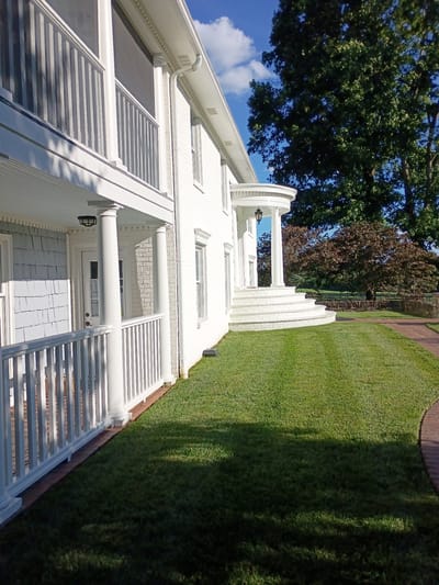 A white two-story brick house with a wraparound porch is seen from a low angle on a sunny day. The house has white railings and columns, and a rounded entrance with a staircase. A large tree with green leaves is visible in the background. The lawn is well-maintained and there is a brick walkway. The scene is located in Lexington, United States.
