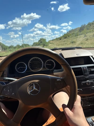 The image is taken from the driver's perspective inside a Mercedes-Benz car, likely in Sotirya, Bulgaria, on a sunny day. The focus is on the steering wheel, dashboard, and the view through the windshield. The steering wheel is a tan leather, with a Mercedes-Benz emblem in the center. To the left of the steering wheel is the turn signal lever, and on the right, a hand is visible gripping the wheel. The dashboard displays the car's instruments, including a speedometer showing 61 km/h, and other gauges. The car's interior appears to be well-maintained. Through the windshield, a road lined with green trees and rolling hills can be seen under a bright blue sky with scattered white clouds. The environment suggests a rural or semi-rural setting.