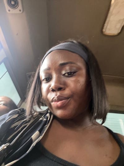 A close-up, vertically oriented selfie shows a Black woman with dark hair, wearing a black tank top and a grey headband. She is looking directly at the camera with a slight smirk. Her skin appears smooth and her eyes are open. She is holding a patterned bag with black and cream stripes. The background is dimly lit and appears to be the interior of a vehicle, possibly a bus, with a beige-colored panel visible in the upper left corner and a circular vent on the left side. The lighting suggests it might be daytime. The image is slightly blurry, contributing to a casual and candid feel. There is no visible text in the image. The subject's expression is neutral to slightly playful.