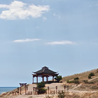 This is a photograph of a traditional East Asian-style gazebo situated on a grassy hillside overlooking the sea. The gazebo is dark red and black, with a tiered roof and open sides supported by pillars. In the foreground, there are red Japanese torii gates and lanterns, as well as low decorative fences. The hillside is covered in dry, brown grass with scattered green bushes and trees. The sea is visible on the left side of the image, with a clear blue sky above. The location is the Chinese Garden of Gardens in Sudak, Crimea, Ukraine.