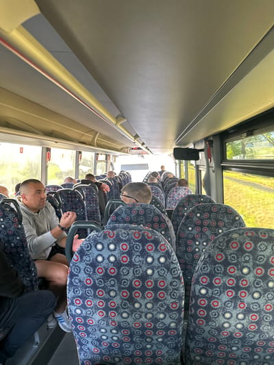 The image depicts the interior of a bus, likely en route to or from Marcinowice, Poland, based on its context. The perspective is from a passenger seated on the right side, looking forward and slightly to the left. The aisle is visible, lined with seats upholstered in a patterned fabric of blue, grey, red, and white circles. Several passengers are seated, with most facing forward.

In the foreground, on the left, a man with a shaved head and a grey hoodie is speaking, gesturing with his hands. He is seated next to a person whose legs and lower torso are visible, wearing dark trousers and athletic shoes. To his right, in the row ahead and slightly further back, another man with glasses is visible from the back of his head. Further down the aisle, other passengers are seated in varying degrees of visibility.

The bus interior features overhead compartments and lighting. Large windows run along the right side, offering a view of a green, grassy area with trees and sunlight filtering through. The overall lighting within the bus is natural, coming from the windows, and appears to be daytime. The atmosphere seems casual, with passengers engaged in conversation or simply traveling.
