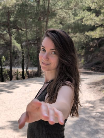 A woman with long, dark brown hair and green eyes smiles at the camera while extending her right hand forward. She is wearing a dark green, knitted top and a silver necklace. She is outdoors in a wooded area with pine trees and a dirt path. The location is Dzhankhot, Russia. The lighting suggests it is daytime.