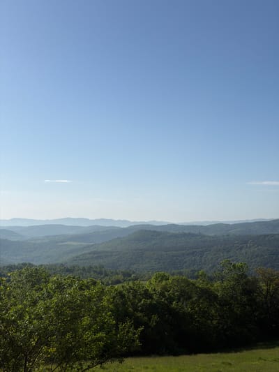 The image shows a landscape with a clear blue sky taking up a large portion of the left side. A thin, white cloud is visible in the sky. On the right side, a range of mountains and hills covered in green vegetation is displayed. The foreground consists of lush green grass and trees. The image does not depict anything that implies it is located in the city of Novogortsi, Bulgaria.