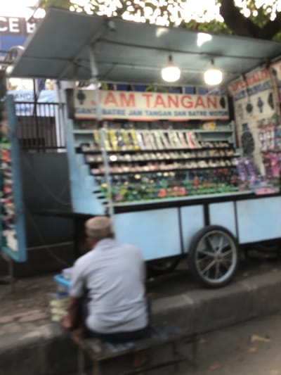The image depicts a street scene in Sumokali, East Java. A person wearing a white shirt and a head covering is visible, appearing to be engaged in some activity, possibly related to a stall or shop. The stall itself is filled with various items, suggesting it might be a retail establishment. There is a prominent vertical sign with red and white text that reads "JAM TANGAN" which translates to "Watches." Other signs with unreadable text are also present. In the background, a bicycle wheel is visible, and the overall setting appears to be outdoors, likely on a street or sidewalk. The lighting suggests it could be daytime. The image is somewhat blurry, making it difficult to discern finer details of the objects and the surrounding environment.