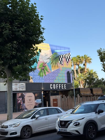 This image depicts the exterior of a coffee shop in Pla Parcial Platja, Spain. A large tree with dark green leaves partially obscures the view of the building's facade, which is covered in a colorful, geometric mural featuring abstract shapes, palm fronds, and coffee beans. The words "COFFEE" are prominently displayed in large white letters across a black awning. Below the awning, a shop window displays an advertisement for "D'ORIGEN COFFEE ROASTERS" celebrating their "10 ANIVERSARIO," featuring an image of a man brewing coffee and a hand holding a blue cup. Two white cars are parked on the street in front of the coffee shop. The car closer to the viewer is a Nissan Juke with license plate "4978 NKS". The car further back is a white Citroën with license plate "7429 JCK". In the background, several palm trees and other greenery are visible under a clear blue sky.
