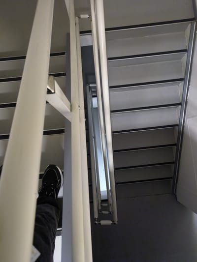 The image shows a person's left foot and lower leg wearing a black sneaker and dark pants, standing on a metal staircase. The staircase has white vertical railings and grey steps with black anti-slip strips. The viewpoint is from above and slightly to the side, looking down the stairwell. The environment appears to be indoors, possibly a public building or apartment complex, given the utilitarian design of the stairs. There is no visible information in the image that would place this scene specifically in Қосшағыл ауылдық округі, Kazakhstan.