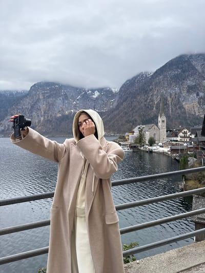 An individual stands outdoors on an elevated platform, positioned behind a dark gray metal railing, in the village of Hallstatt, Austria. The person, wearing a long, light beige overcoat over a cream-colored hooded sweatshirt and pants, holds a black digital camera in their extended right hand, angled towards themselves as if taking a selfie or recording video. Their left hand is raised near their face. Behind the individual, the dark, rippled waters of Hallstätter See extend to a narrow strip of the village, dominated by a light-colored stone church featuring a tall, pointed spire and clock tower. Numerous traditional buildings with dark roofs are clustered along the waterfront and ascending the lower mountain slopes. Several small boats and at least one larger white vessel are visible on the lake near the village. Steep, rugged mountains, partially covered in bare trees and patches of snow near their peaks, rise directly from the lake on the opposite shore. The sky is overcast and gray, with low clouds obscuring some mountain tops.