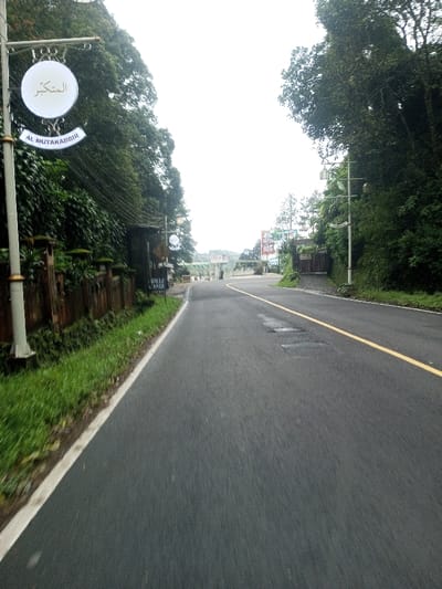 This image displays a view along an asphalt road in Deothang, Bhutan, under an overcast sky. The road, appearing to be a single lane in this perspective, features a solid yellow center line and a white edge line on the left. Patches of wear are visible on the road surface.
On the immediate left, a light grey pole with decorative golden accents supports a circular white sign bearing Arabic script ("المتكبر") and the English transliteration "AL MUTAKABBIR." Below this, a partially visible sign reads "VILLE AMAR." A dark wooden fence with stone-capped posts runs along the left side of the road, bordered by short green grass. Beyond the fence, dense green foliage and trees line the road.
On the right side, lush green vegetation, primarily trees and bushes, covers a rising embankment. Further down the road, a paved turn-off leads to a dark wooden gate. In the mid-ground, an elevated structure, possibly an overpass or bridge, spans the road. Multiple colorful billboards are visible beyond this structure in the distance. No individuals are present in the scene.