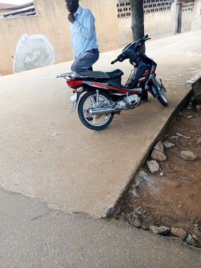 In Egbedi, Nigeria, a man wearing a light blue button-down shirt and dark trousers kneels on a concrete surface next to a black and red motorcycle. The motorcycle has a chrome exhaust pipe and a license plate. The ground is a mix of concrete and dirt, with some rocks and debris visible. In the background, a light-colored wall with a textured patch and a series of square openings can be seen, along with a tree trunk. The scene appears to be outdoors, possibly in a public area or residential compound.
