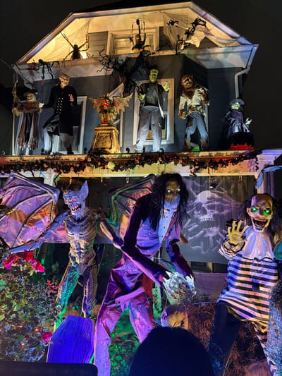 An external view of a multi-story residential building located in Weehawken, United States, displays an extensive Halloween decoration setup at night. The house features light-colored siding and is illuminated by various colored lights, including white, green, and purple.
The attic gable area is adorned with large spider props, white translucent web material, dark skeletal branches, and a smaller suspended figure near the central window.
On the second-story balcony, arranged from left to right, are several full-sized figures: a draped scarecrow-like entity; a male figure resembling a vampire or formal ghoul in a dark coat; a central pedestal supporting a winged gargoyle prop; a green-skinned Frankenstein's monster figure with outstretched hands; a standing werewolf figure in torn clothing; and a seated witch figure in a black cloak. The balcony railing is draped with dark foliage garland interspersed with multi-colored string lights.
The ground-level or lower porch area features additional large figures and props. On the left, a skeletal bat-creature with large wings and prominent ribs stands amidst green foliage and red flowers. A blue coffin-shaped tombstone prop inscribed "ANNA KODEN" is partially visible. Centrally positioned is a tall, slender figure with long dark hair, glowing white eyes, and a tattered suit illuminated in purple light, holding a spiky prop. To its right, a clown-like figure with glowing green eyes, a wide grin, and a white and black striped shirt extends a hand. A large backdrop behind these figures depicts a skull surrounded by reaching skeletal hands. No actual people are visible; all subjects are static decorative props.