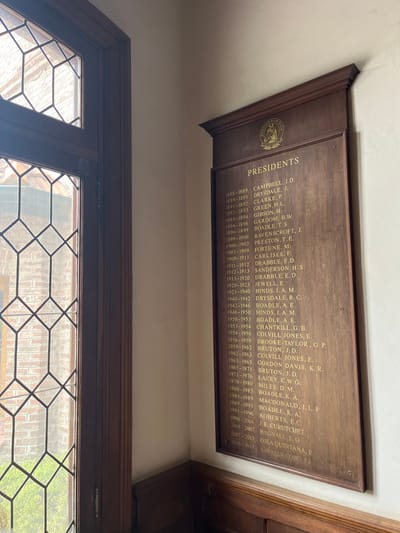 The image displays a long, dark wooden plaque mounted on a light-colored wall, with a leaded glass window visible below it. The image is oriented with the plaque appearing on its side, but the text indicates it's meant to be read horizontally.

The plaque itself is made of a rich, dark brown wood, possibly mahogany or oak, with a slightly glossy finish. It has a decorative molded edge along its top and bottom. On the left side of the plaque, there is a circular golden emblem or crest, though the details of the design are difficult to discern clearly. To the right of the emblem, the word "PRESIDENTS" is inscribed in large, golden, sans-serif capital letters.

Below "PRESIDENTS," a detailed list of names, each accompanied by a date range, is inscribed in smaller, golden capital letters, formatted in two columns. The list spans from 1888-1889 up to 2013-2015. The entries are:

*   1888-1889 CAMPBELL, J. D.
*   1889-1891 DRYSDALE, J.
*   1891-1892 CLARKE, P.
*   1892-1893 GREEN, H.L.
*   1893-1894 GIBSON, H.
*   1894-1896 GARDOM, B.W.
*   1896-1899 BOADLE, T.S.
*   1899-1900 RAVENSCROFT, J.
*   1900-1903 PRESTON, T.E.
*   1903-1909 FORTUNE, M.
*   1909-1911 CARLISLE, F.
*   1911-1912 DRABBLE, E.D.
*   1912-1913 SANDERSON, H. S.
*   1913-1920 DRABBLE, E. D.
*   1920-1923 JEWELL, E.
*   1923-1940 HINDS, J. A. M.
*   1940-1942 DRYSDALE, R. C.
*   1942-1946 BOADLE, A. E.
*   1946-1950 HINDS, J. A. M.
*   1950-1953 BOADLE, A. E.
*   1953-1954 CHANTRILL, G. B.
*   1954-1956 COLVILL JONES, E.
*   1956-1959 BROOKE TAYLOR, G. P.
*   1959-1962 BRUTON, J. D.
*   1962-1963 COLVILL JONES, E.
*   1963-1965 GORDON DAVIS, K. R.
*   1965-1971 BRUTON, J. D.
*   1971-1976 LACEY, C.W.G.
*   1976-1980 MILES, D. M.
*   1980-1985 BOADLE, K. A.
*   1985-1989 MACDONALD, I. L. P
*   1989-1991 BOADLE, K. A.
*   1991-1996 ROBERTS, E.C.
*   1996-2004 J. E. CURUTCHET
*   2004-2007 BAGNALL EO
*   2007-2013 QJEA QUINTANA, E
*   2013-2015 LAVALLE COBO, FJ

The wall behind the plaque is painted a plain, light cream or off-white color. Below the plaque, a window with a dark wooden frame is visible. The window features multiple small panes of glass separated by dark lead cames, forming a hexagonal grid pattern. Through the window, blurred green foliage and what appears to be a light-colored brick structure can be seen, suggesting an outdoor view.

The lighting in the image appears to be natural, coming from the window, casting a soft, even light on the plaque and wall. The photo is taken from a slightly low and angled perspective, looking up towards the plaque and across towards the window.