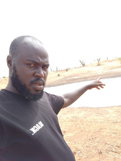 A man with dark skin, a beard, and a bald head is standing outdoors in Mulke Barikin Sayejo, Nigeria. He is wearing a black t-shirt with the word "BLACK" printed in white. He is pointing his right arm towards a bright white surface in the background, possibly a body of water reflecting light. The landscape consists of sparse vegetation and dry, sandy ground.