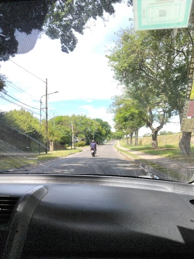 Captured from a car's interior, the image showcases a straight road stretching into the distance under a bright, slightly cloudy sky. A lone motorcyclist rides ahead. Lush green trees line both sides of the road, creating a canopy effect in some areas. Utility poles with wires run along the left side, adding to the suburban atmosphere. The car's dashboard dominates the foreground, providing a clear point of view from within the vehicle.