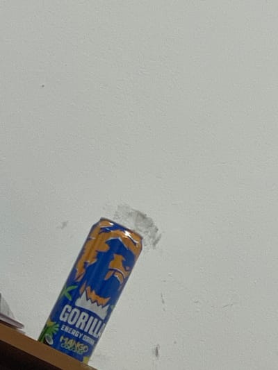 A partially visible blue and orange can of "Gorilla Energy Drink Mango Coconut" is leaning against a textured white wall in Рахат ауылдық әкімдігі, Kazakhstan. The can is angled to the left and positioned in the lower center of the frame. The wall appears to have some imperfections and dust. A wooden surface is visible at the bottom left of the image, beneath the can. The scene is indoors and appears to be in a casual setting, possibly a home or an office.
