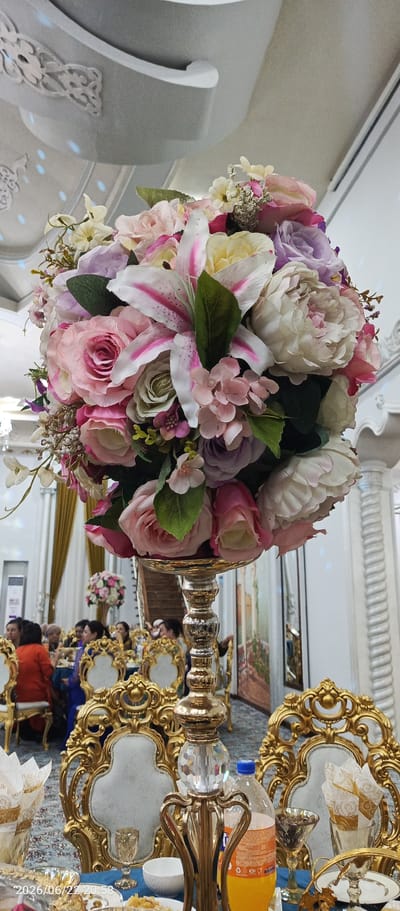 The image depicts a floral centerpiece, likely for a formal event, prominently displayed on a table. The centerpiece is a large, round arrangement of artificial flowers in various shades of pink, white, and lavender, featuring prominent lilies and roses. It is supported by an ornate, gold-colored pedestal with crystal accents. The table itself is draped with a blue tablecloth and set with golden-trimmed chairs, some of which are occupied by people. In the background, other tables and decorative elements of a banquet hall are visible, including golden curtains, pillars, and wall decor. The overall atmosphere suggests a celebratory or formal gathering. The timestamp "2026/06/22 20:58" is visible in the lower left corner of the image. While the image is specified to be from Vabkent, Uzbekistan, there are no specific visual cues within the image that directly confirm this location.