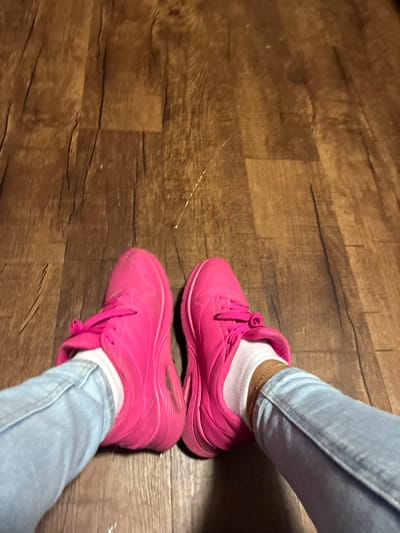 A pair of bright pink athletic shoes with white socks are shown from a first-person perspective, with the wearer's light-wash denim jeans visible at the bottom. The shoes are laced and appear to be in good condition. A delicate gold chain anklet is visible on the right ankle. The shoes and legs are resting on a wooden floor with a visible wood grain pattern. The image provides no indication of location in Dallas, United States of America.