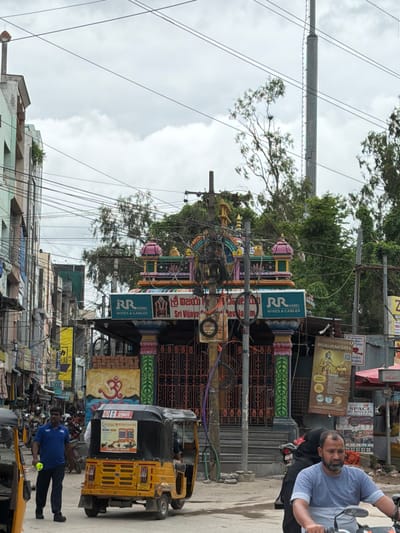 The image captures a street scene in Shamshabad, India, under an overcast sky. The foreground features a bright yellow auto-rickshaw, facing right, with a man in a blue t-shirt walking beside it to the left, holding a yellow object. Behind the auto-rickshaw, a man wearing a light blue t-shirt and a beard rides a motorcycle, looking forward. To the right of the auto-rickshaw, a woman in a black hijab is partially visible.
Dominating the center of the image is a colorful, ornate structure, likely a Hindu temple or shrine, with vibrant pillars, intricate carvings, and tiered rooftops. A sign above the entrance reads "RR KABEL WIRES & CABLES" in English, and also in a regional script. Another sign above that displays "శ్రీ విజయ" and "Sri Vijaya". To the left of this structure, a large yellow sign with Telugu script and an "Om" symbol is visible on the wall. Further to the left, buildings with multiple floors and balconies line the street, with numerous electrical wires crisscrossing the scene. Several other vehicles, including motorcycles and auto-rickshaws, are present further down the street, suggesting a bustling urban environment. Advertisements and signs are plastered on various surfaces. The overall impression is of a busy street in an Indian city with a strong religious presence.