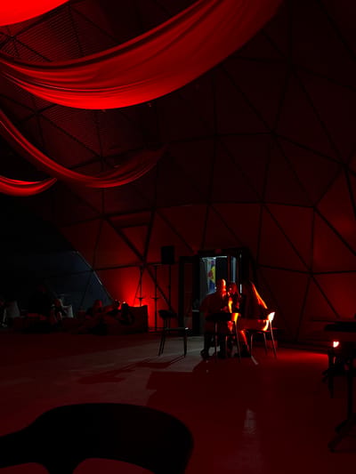 The image depicts an interior scene illuminated by red lighting. In the foreground, a dark, abstract shape is visible on the floor. In the midground, several people are seated on a large, low sofa or cushion area, with some interacting. Further back, a group of three people are seated at tables, with two facing each other and one turned away. Behind them, an open doorway reveals a glimpse of what appears to be a bar area with Red Bull branding. The environment appears to be a dome-like structure with visible metal framework. Red fabric drapes from the ceiling. The overall impression is that of a dimly lit, atmospheric gathering or event space. This scene is located in Žemaitkiemis, Lithuania.