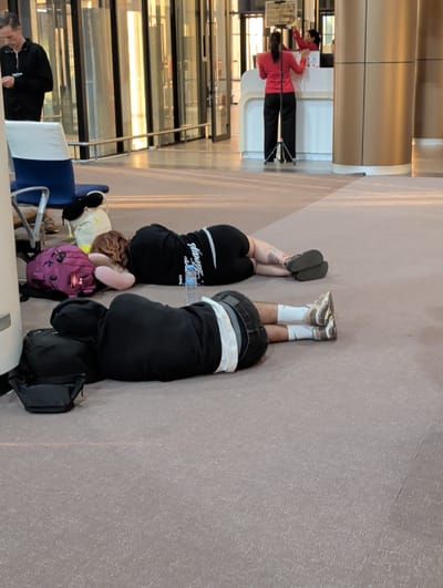 The image shows two people lying on the floor, with their legs up in the air. They appear to be in an airport terminal, with polished floors and large windows in the background. The lighting suggests it is daytime. One person is wearing black shorts and white socks with athletic shoes, while the other is wearing black leggings and a black shirt with white text that reads "Always". There are several pieces of luggage around them, including a purple backpack and black bags. In the background, other people can be seen standing at a counter, some wearing red shirts. The overall atmosphere appears to be one of waiting or resting.