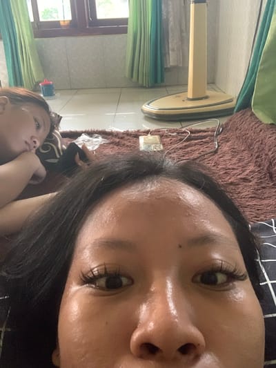 Two people are lying on a brown furry rug in what appears to be a residential interior. The person in the foreground is facing the camera, with their face taking up most of the frame. Their eyes are wide open, and they have long, dark eyelashes. The person in the background is lying on their side, with their head resting on a pillow. They are looking away from the camera. A floor fan is visible in the background, along with green curtains and a window. There is also a small blue cup with a red lid on the floor. The scene suggests a casual, indoor setting. It's not possible to determine the exact location in Jajag, Indonesia based solely on the visual information provided.
