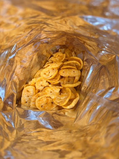 The image shows a close-up, overhead view of a pile of orange-colored, circular, savory snack chips inside a shiny, crinkled, golden-colored bag. The bag appears to be made of a metallic material, possibly foil, and is partially open, revealing the chips within. The chips have a slightly irregular texture and are dusted with seasoning. The lighting in the image is bright, reflecting off the bag and highlighting the texture of the chips. The background is out of focus and consists of the reflective interior of the bag. There are no people or signs of outdoor environment visible. This scene could be anywhere, but it is unlikely to be a specific location within Tēraudi, Latvia without further context.