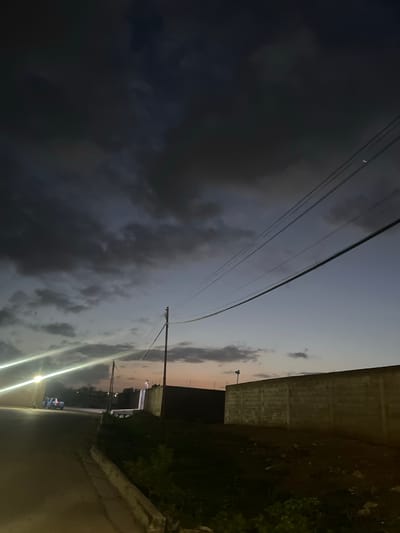 A nighttime, low-angle photograph captures a street scene in La Cruz del Pastel, Venezuela, under a dark, cloudy sky. In the foreground, a paved road curves to the left, bordered by a concrete curb. To the right, a rough, unpaved area is visible, sloping upwards towards a tall, light-colored brick wall. The wall has a rough, unfinished appearance, with visible mortar lines between the blocks. In the distance, silhouetted trees and low-rise buildings are partially obscured by darkness. A street light illuminates the left side of the road, casting bright streaks of light. A light-colored pickup truck is parked on the side of the road, its headlights on. In the sky, a utility pole with several wires is visible, stretching across the frame. The clouds are dark and heavy, with some lighter patches suggesting residual daylight or distant illumination. The overall atmosphere is dim and somewhat moody, characteristic of a twilight or early night setting.