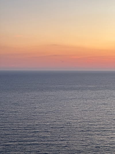 A serene seascape is depicted, with the ocean filling the lower two-thirds of the frame and the sky occupying the upper third. The ocean's surface is calm, exhibiting a gentle ripple pattern that stretches to the horizon. The water's color is a deep, muted blue-gray.
The sky above transitions from a soft, pale gray at the very top to a gradient of warm hues. The colors range from a pale yellow-orange near the horizon, blending into soft pinks and peachy tones, and finally fading into the pale gray. A few faint, wispy clouds are present, subtly catching the light.
The image captures the tranquil atmosphere of a coastal scene, likely at dawn or dusk, in the vicinity of Glavatičići, Montenegro. There are no people, specific objects, or actions visible in the frame; the focus is solely on the natural expanse of the sea and sky.