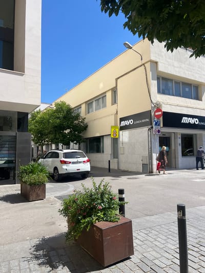 The image depicts a street view in Granollers, Spain, specifically in front of the "MAYO CLÍNICA DENTAL" building. The building is multi-storied and cream-colored, and has a second sign that reads "MAVO CLÍNICA". A street lamp is affixed to the building's facade. Street signs indicating "Zona escolar" (school zone) and speed limits are posted near the clinic. A white car is parked along the curb. A few people are walking on the sidewalk near the clinic's entrance. There are bollards and planters with greenery are lining the sidewalk. The sky is clear blue. The architecture is modern.