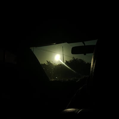 The image depicts a view through a car windshield during a bright sunset. The sun, a luminous orb, is positioned centrally in the frame, casting a strong glare and a visible light beam. A utility pole and power lines are silhouetted against the sky. Trees and foliage form a dark mass in the background, suggesting a rural or semi-rural setting. The interior of the car is largely in shadow, with a portion of the driver's seat visible in the foreground, its textured surface catching a sliver of light. The scene is devoid of any identifiable people or specific landmarks that would indicate the city of Patek, Indonesia.