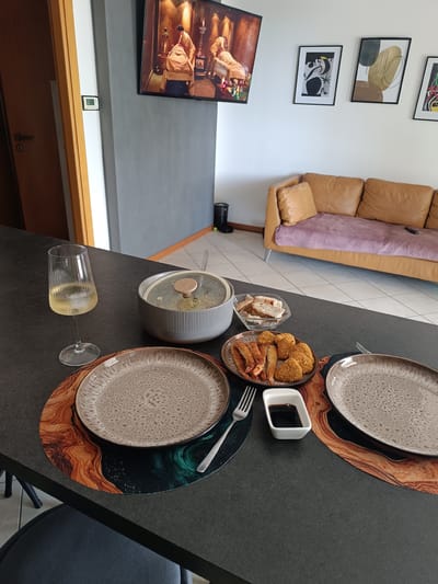 In a modern apartment setting, possibly in Rimini, Italy, a table is set for a meal. The table is dark grey, and on it are two plates, a glass of white wine with ice, a pot with a lid containing food, a bowl of bread slices, a plate of fried food including shrimp and nuggets, and a small dish of soy sauce. A fork rests on one of the placemats. In the background, a large television displays an image, and further beyond, a tan leather sofa is visible with a pink throw. Several framed abstract art pieces hang on the white wall above the sofa. To the left, a partially open wooden door reveals a doorway, and a white wall with a digital thermostat. The floor is tiled.